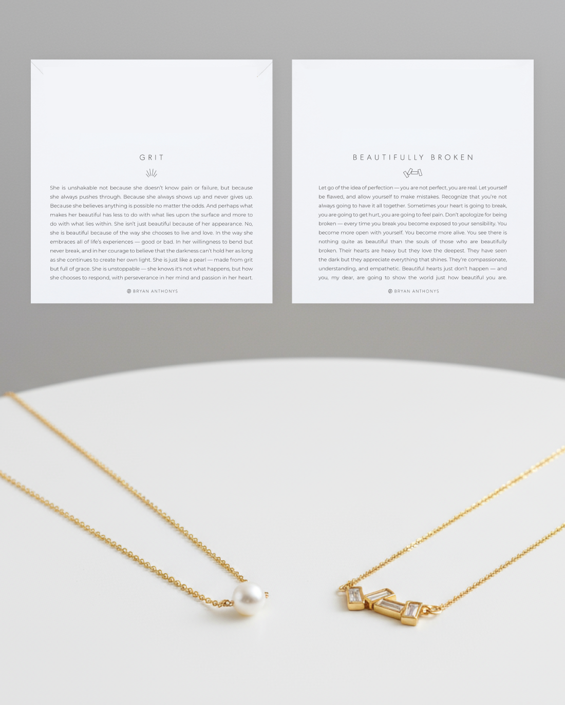 Two white cards, one titled 'GRIT' (left) and one titled 'BEAUTIFULLY BROKEN' (right), sit above two gold chain necklaces. The left necklace has a single pearl pendant, and the right necklace has a charm featuring three rectangular, clear baguette stones.
