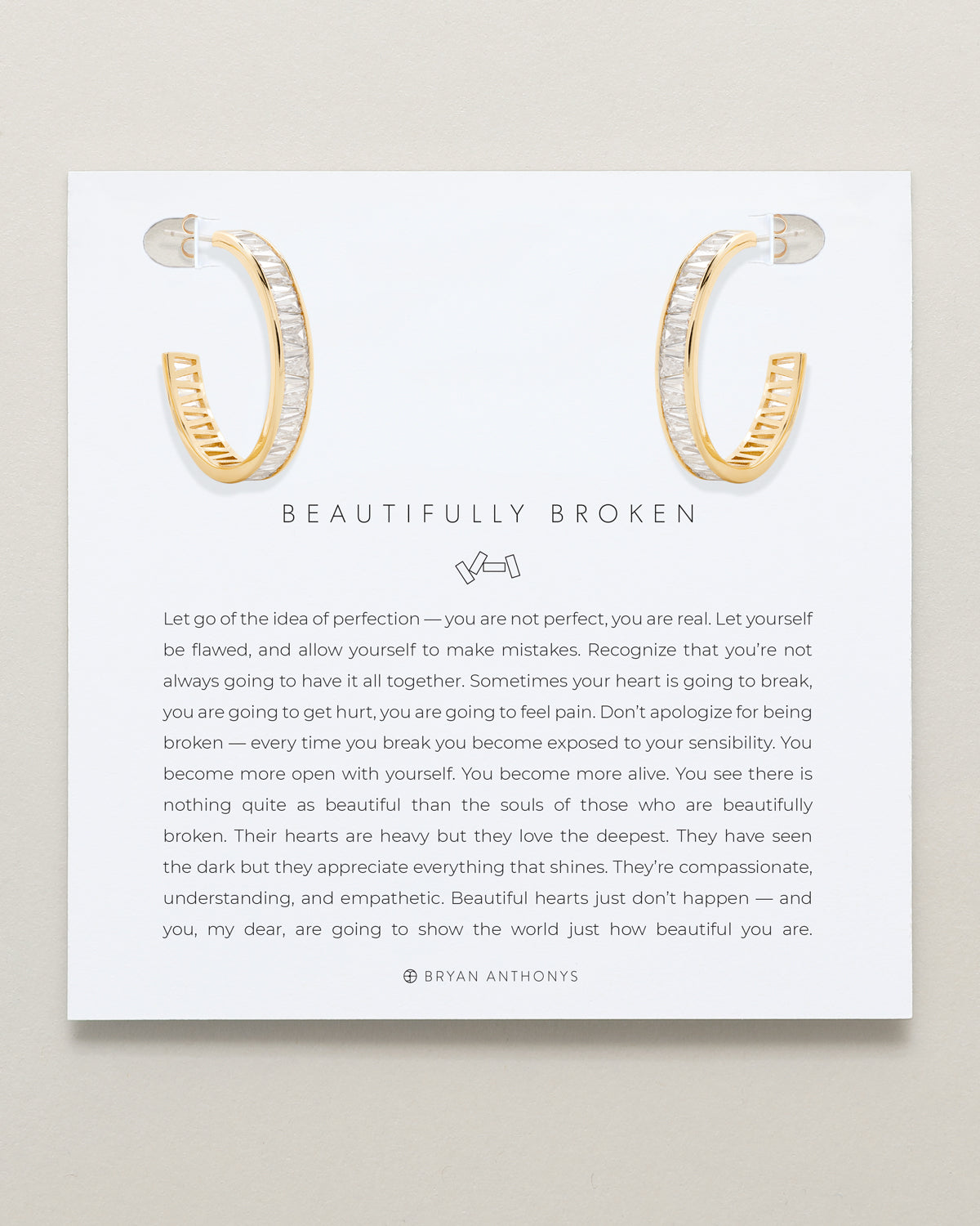 A pair of gold-toned open hoop earrings, featuring diagonally set baguette-cut white stones along the front, are displayed at the top of a white card. The card features the title 'BEAUTIFULLY BROKEN' and a long inspirational message about letting go of perfection.