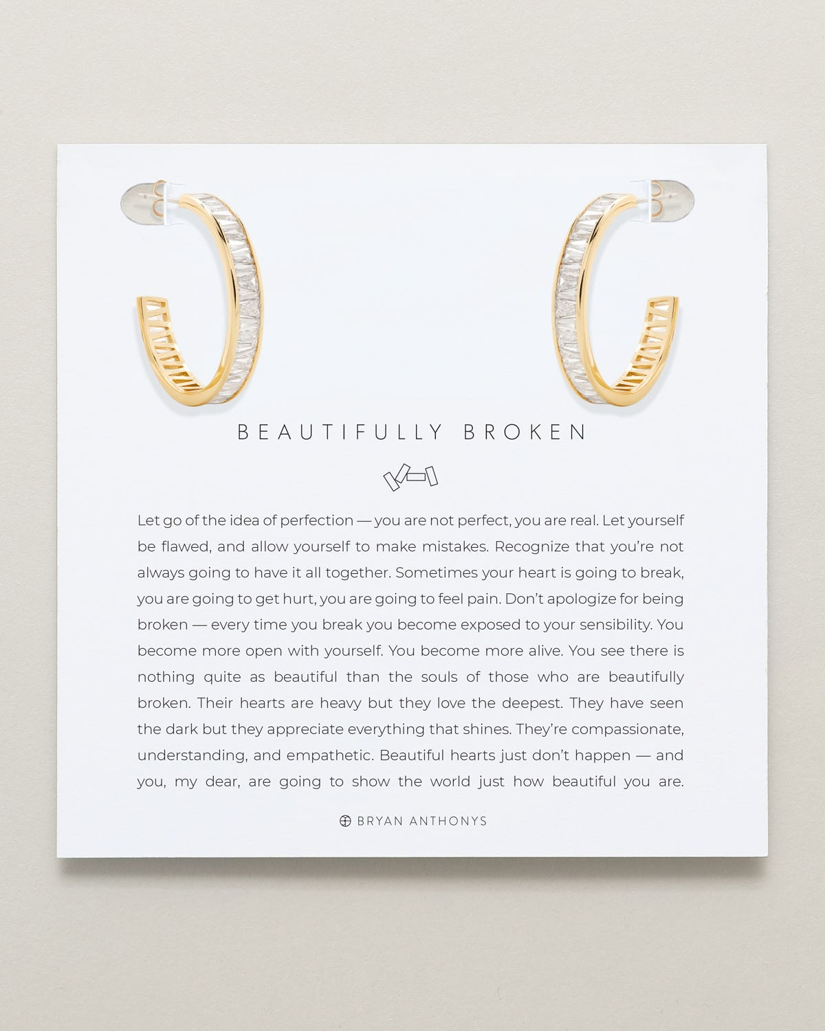 A pair of gold-toned open hoop earrings, featuring diagonally set baguette-cut white stones along the front, are displayed at the top of a white card. The card features the title 'BEAUTIFULLY BROKEN' and a long inspirational message about letting go of perfection.
