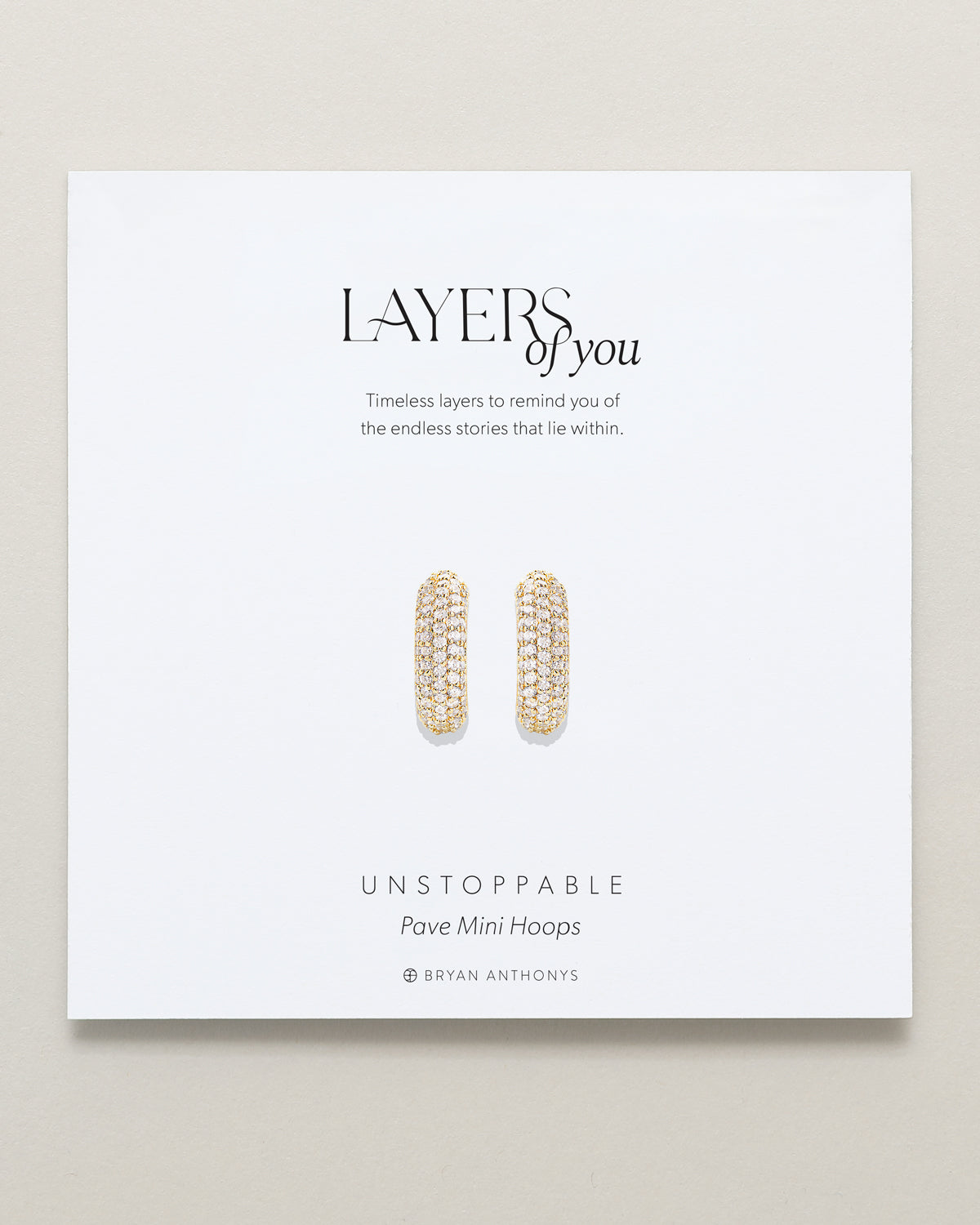 A pair of small gold mini hoop earrings, completely covered in brilliant pave-set stones, are displayed on a white card. The card features the title 'LAYERS of you,' a short inspirational message.