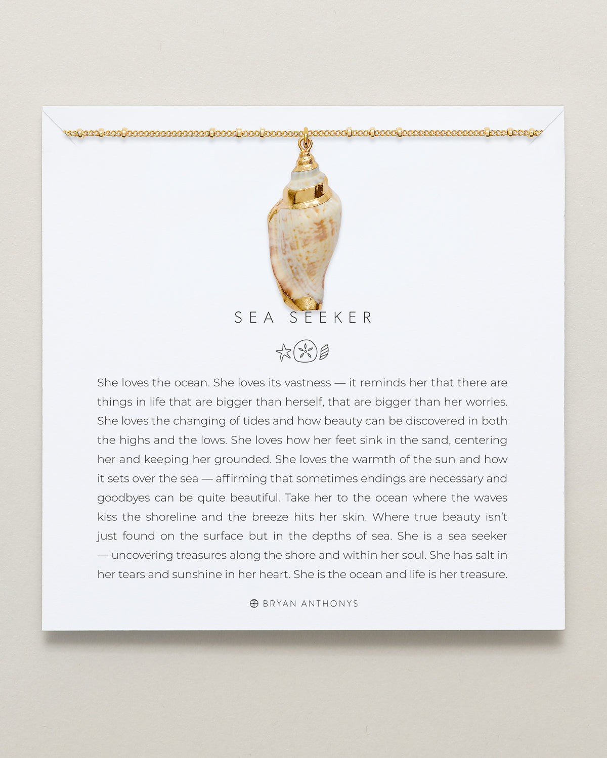 A gold ball chain necklace with a large, polished natural conch shell pendant hangs from the top edge of a white card. The card features the title 'SEA SEEKER' and a long message describing a person who loves the ocean.
