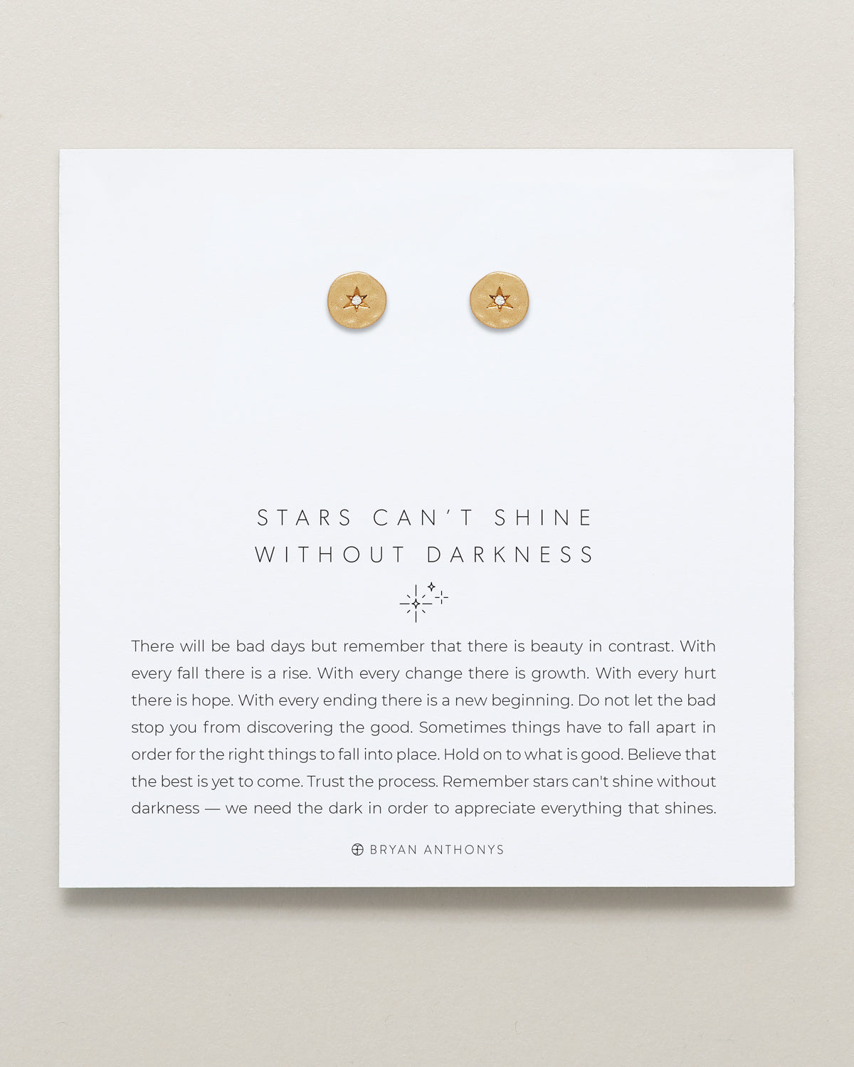 A pair of small, round gold stud earrings with a subtle celestial star design, displayed at the top of a white card. The card features the quote 'STARS CAN'T SHINE WITHOUT DARKNESS' and an encouraging message about finding hope and good after setbacks.