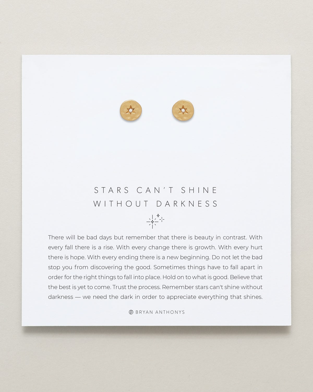 A pair of small, round gold stud earrings with a subtle celestial star design, displayed at the top of a white card. The card features the quote 'STARS CAN'T SHINE WITHOUT DARKNESS' and an encouraging message about finding hope and good after setbacks.