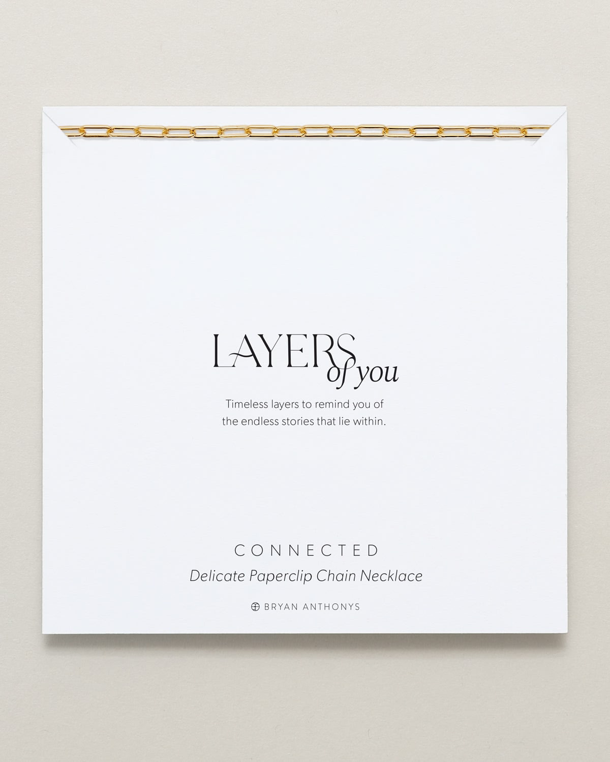 A delicate gold paperclip chain necklace is draped across the top edge of a white card. The card features the title 'LAYERS of you,' a short message about timeless layers.