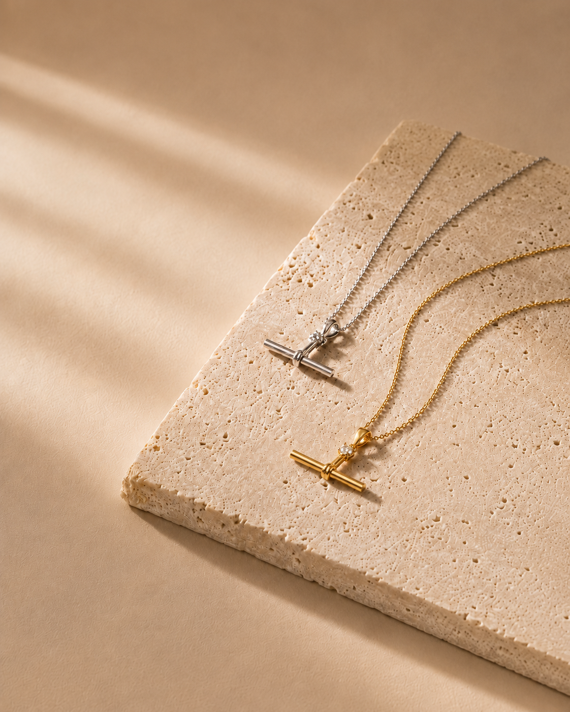Two delicate toggle-bar necklaces, one gold and one silver, displayed on a textured beige stone slab against a warm beige background with soft diagonal shadows.