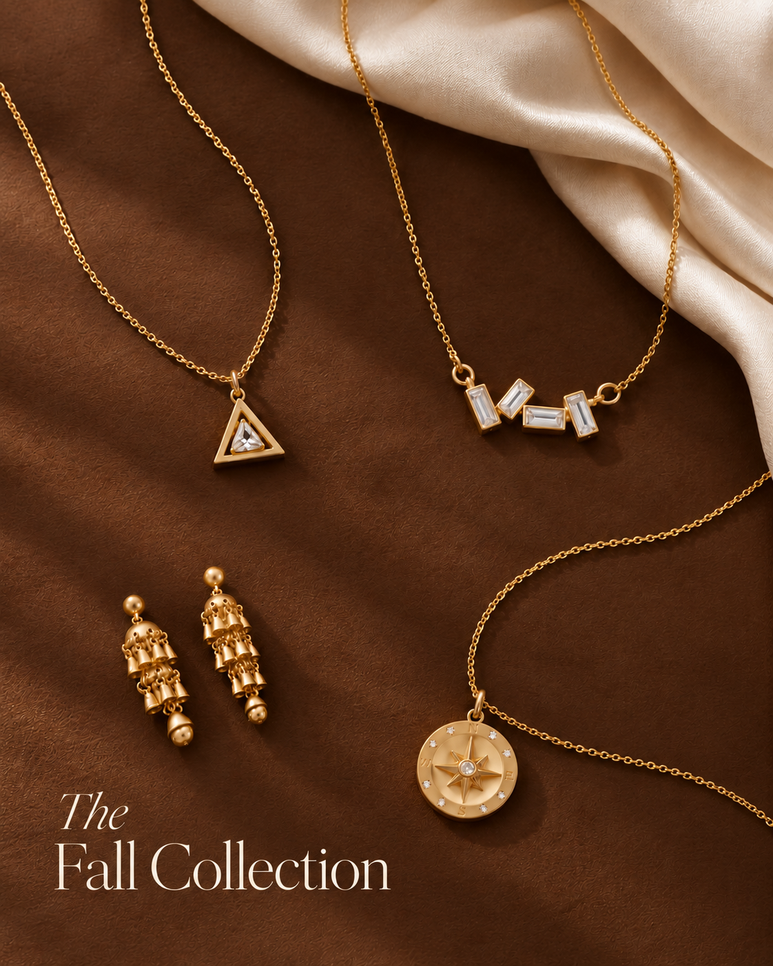 Gold jewelry collection featuring three necklaces and a pair of drop earrings displayed on a brown textured background with draped cream satin. The necklaces include a triangle pendant with a blue stone, a geometric block-letter pendant spelling 'MIL', and a round compass pendant.