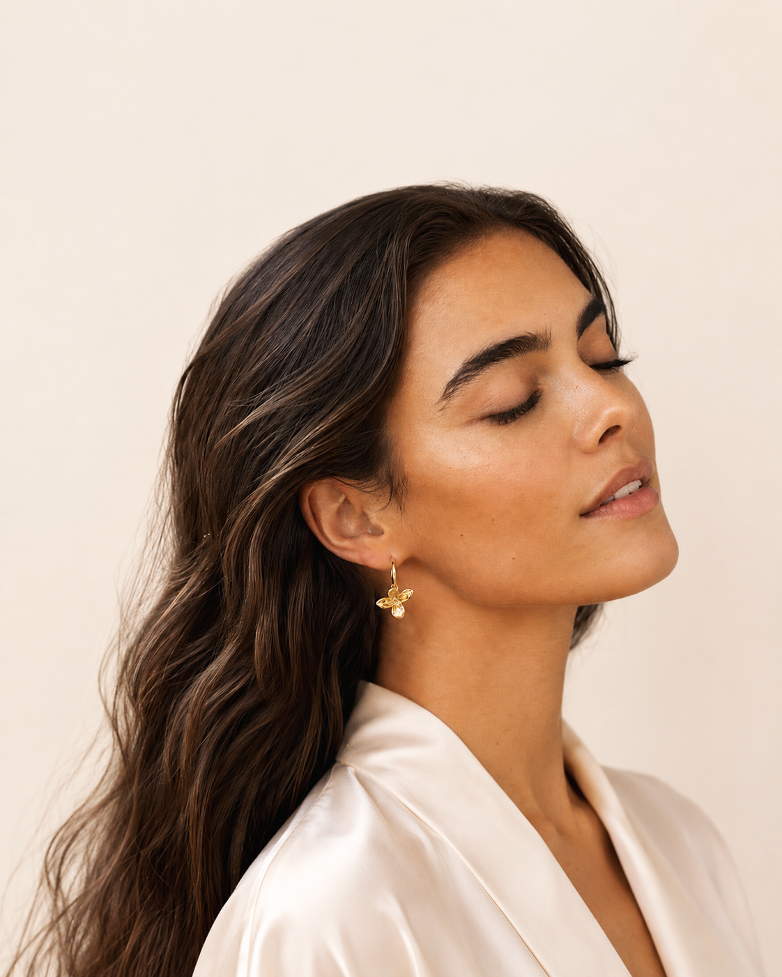 A woman with long dark brown hair is shown in profile wearing a small gold flower-shaped earring. She is wearing a light cream satin top against a plain, warm off-white background.