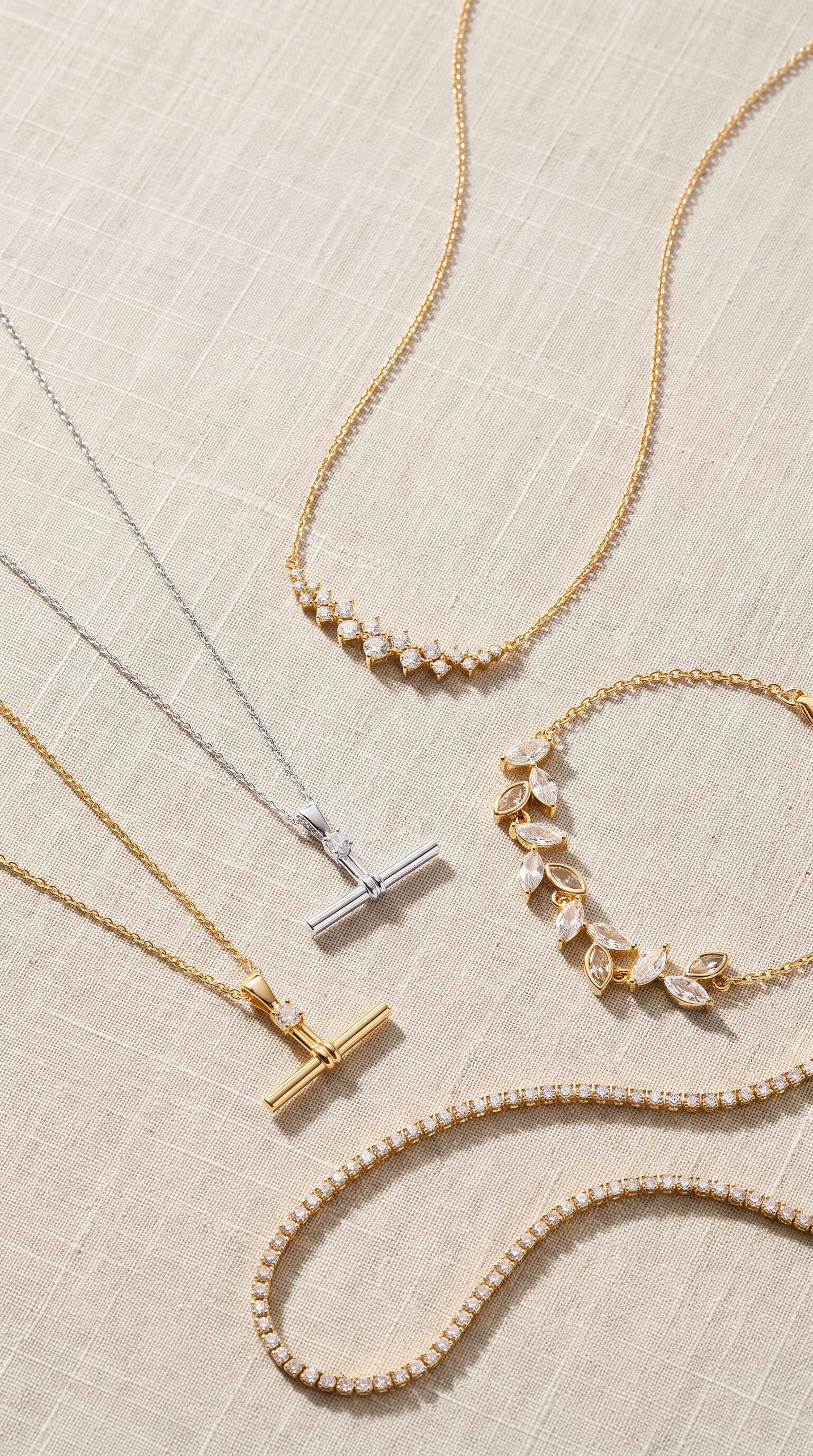 A styled flat-lay of delicate jewelry on a light beige stone surface, featuring gold and silver chains, pearl bead details, and T-bar pendants.